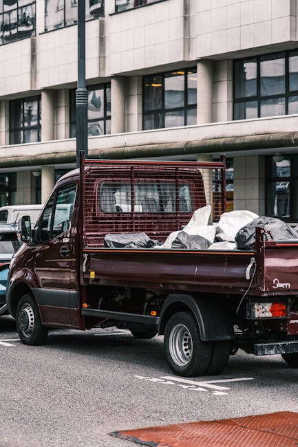 A small burgundy flatbed truck parked on an urban street in front of a modern office building with large glass windows and concrete panels. The truck's cargo area is filled with a variety of white and
